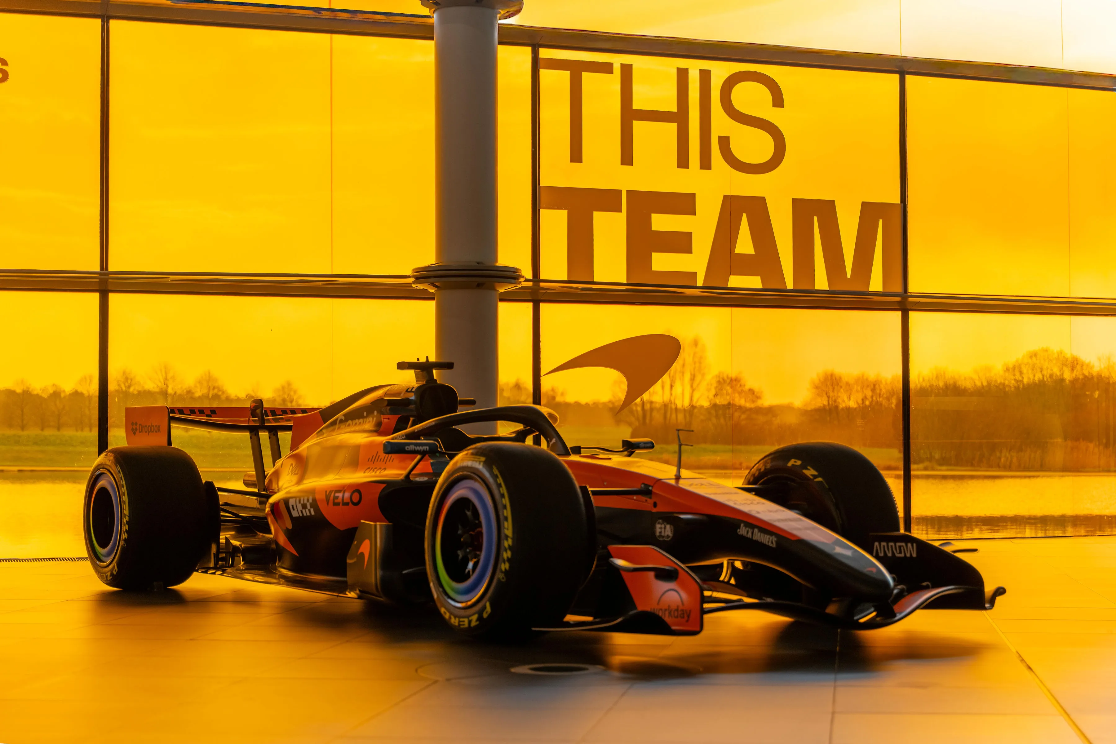 McLaren Formula 1 car backlit by orange evening light through the glass facade of the Technology Centre