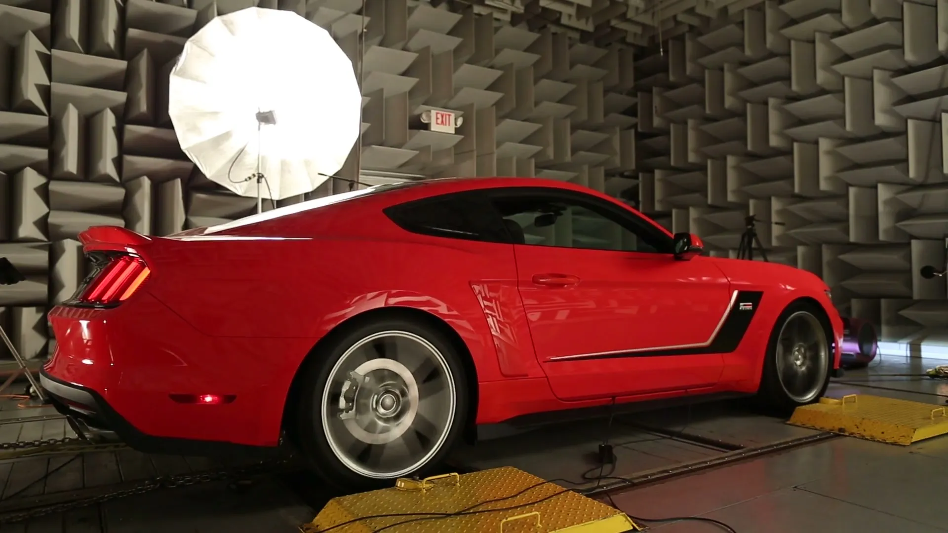 Red ROUSH Mustang on a rolling road inside a semi-anechoic chamber lined with grey acoustic wedges, lit by a single softbox